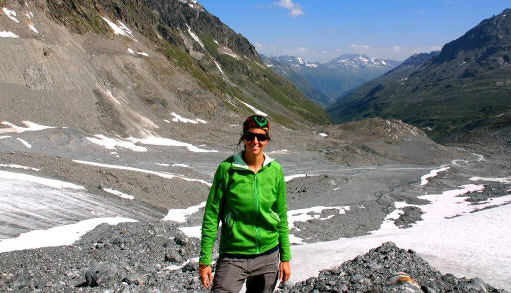 Eine Person in einer grünen Jacke und Sonnenbrille steht in einer felsigen, schneebedeckten Berglandschaft mit fernen Gipfeln unter einem klaren blauen Himmel.