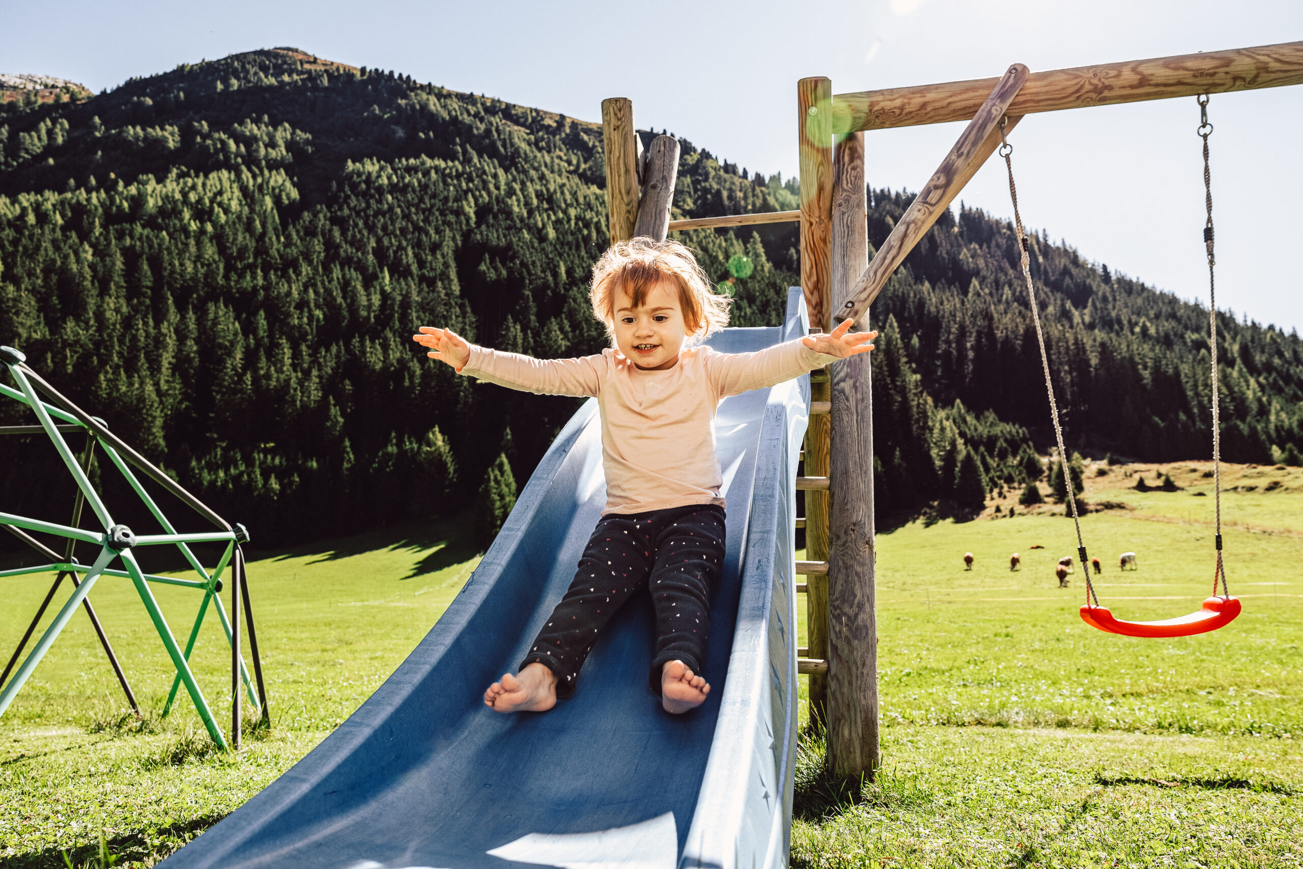 Ein kleines Kind mit braunen Haaren und hellem Hemd rutscht mit ausgestreckten Armen eine blaue Rutsche in einem sonnigen Park in der Nähe der Ferienwohnung Galtür hinunter. Grasflächen, Bäume und Berge bilden die Kulisse, während rechts eine Schaukel und links Spielgeräte stehen.